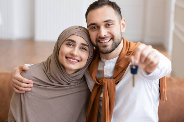 Happy Muslim Family Couple Showing New House Key To Camera Sitting On Sofa Indoors. Own Home, Real Estate Ownership And Housing. Mortgage And Apartment Purchase. Selective Focus